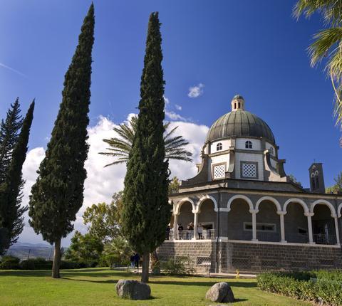 Arched portico surrounding a central dome-topped tower against a blue sky. Tall and thin cyperus trees on a lawn in the forground.
